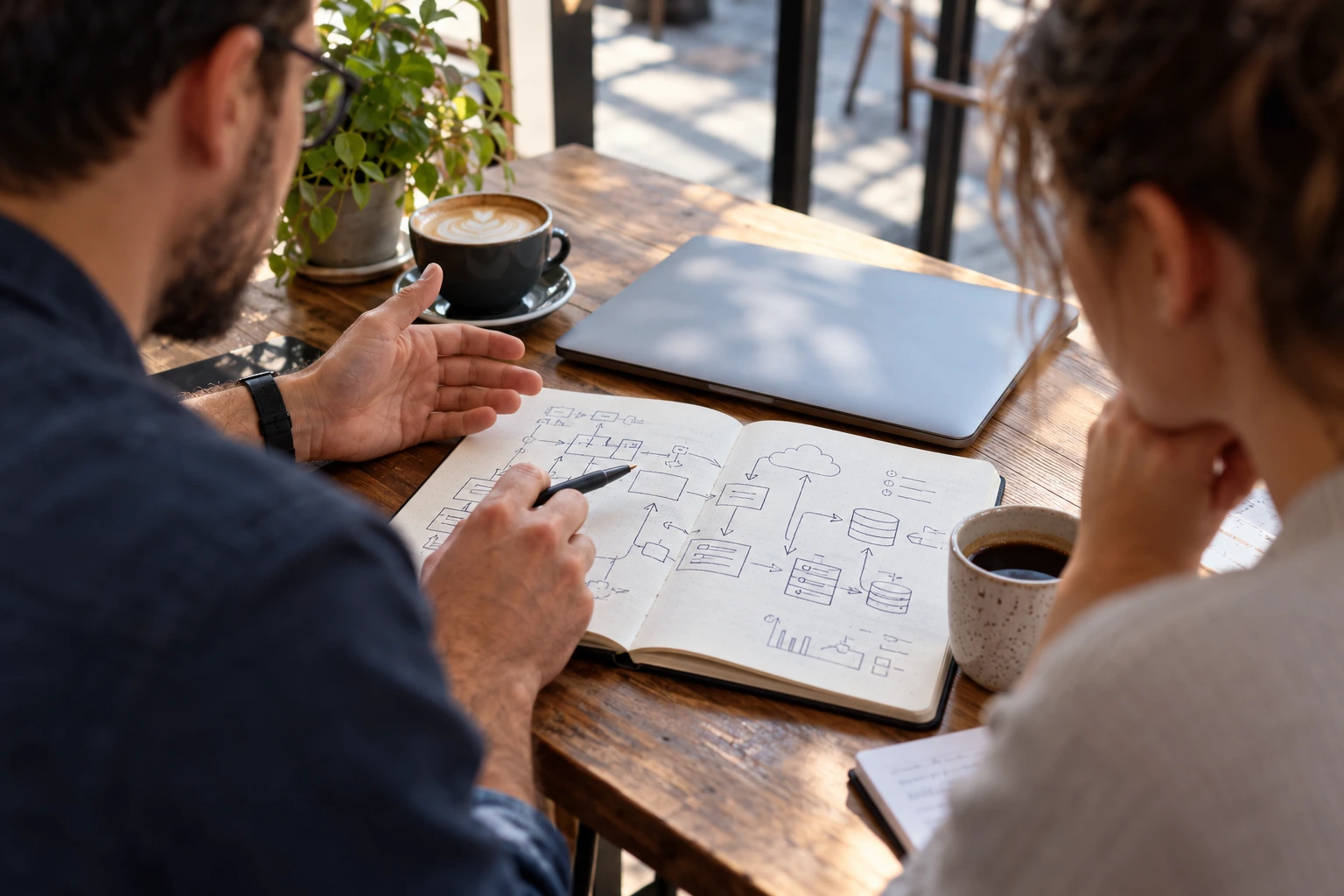 Two colleagues discussing a system sketch in a notebook at a cafe table